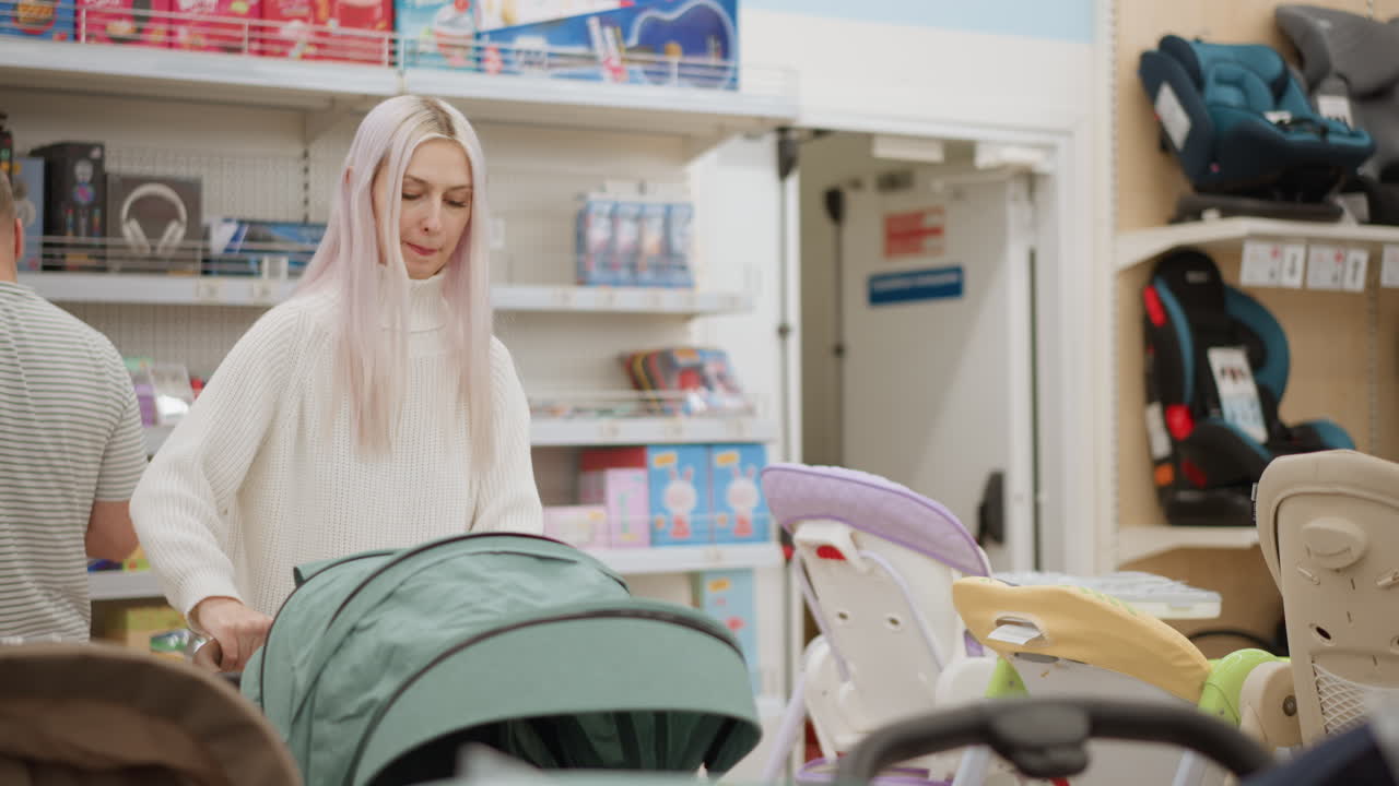Elegant young woman meticulously inspects stroller canopy in bright baby section opening closing cover to check for damage while male shopper browses behind colorful toy aisle display