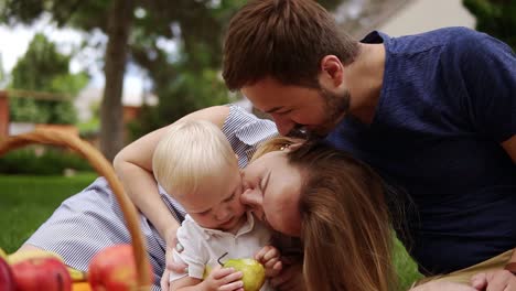 Close-up-of-a-young-parents-sitting-close-to-each-other-with-their-little-son.-Picnic-outdoors-on-a-plaid.-Picnic-basket-with-apples.-Smiling,-happy-family