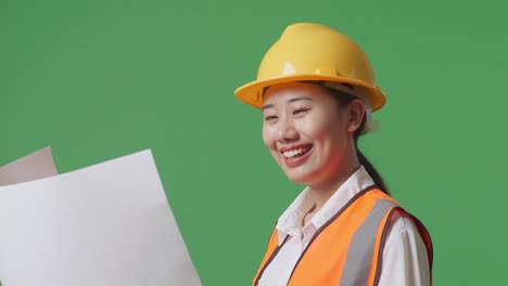 close up side view of asian female engineer with safety helmet looking at blueprint in her hands and looking around while standing in the green screen background studio