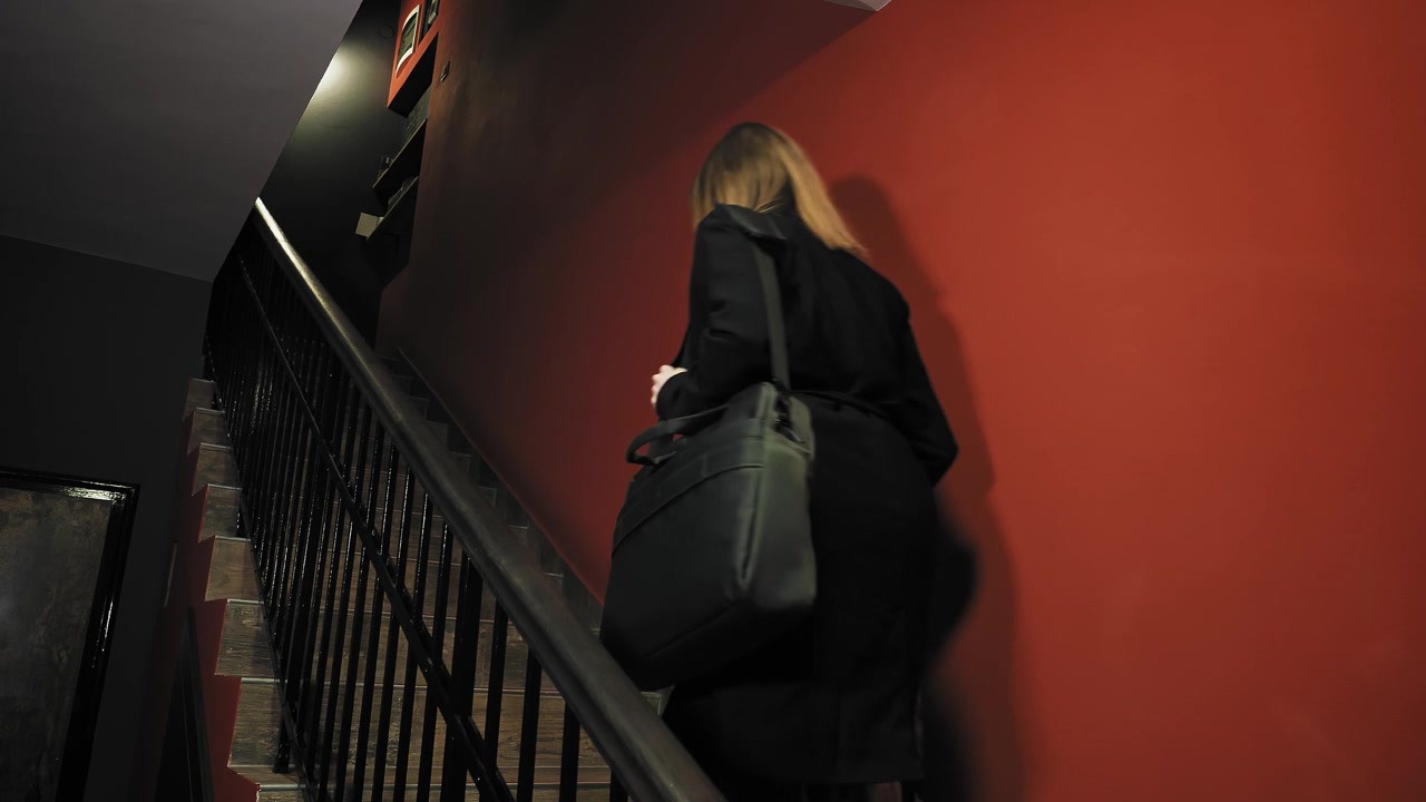 Young woman carries rolling suitcase up a flight of stairs in hotel; red colored hallway walls.