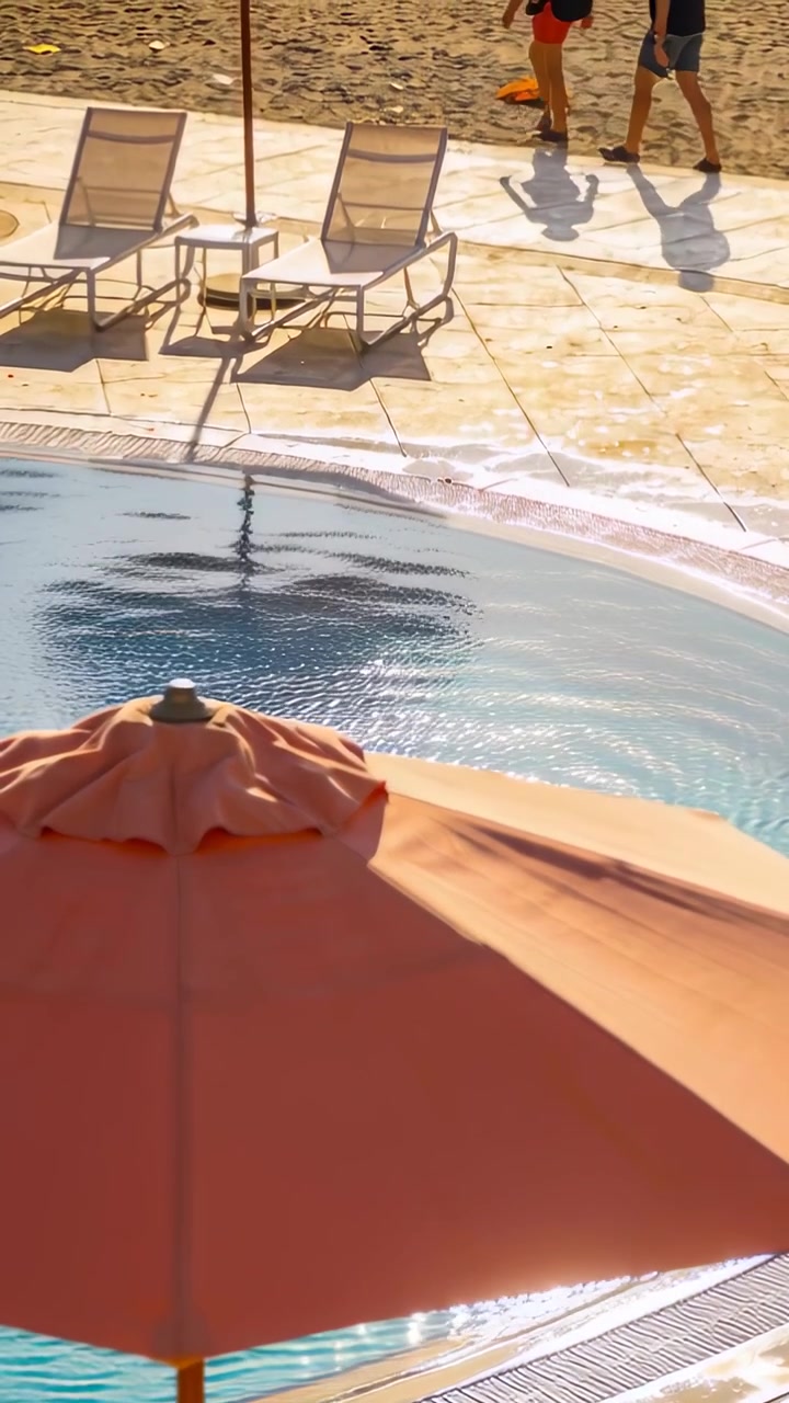 Vertical video: Panning cam showing two adults walking along pool deck toward beach, coral umbrella