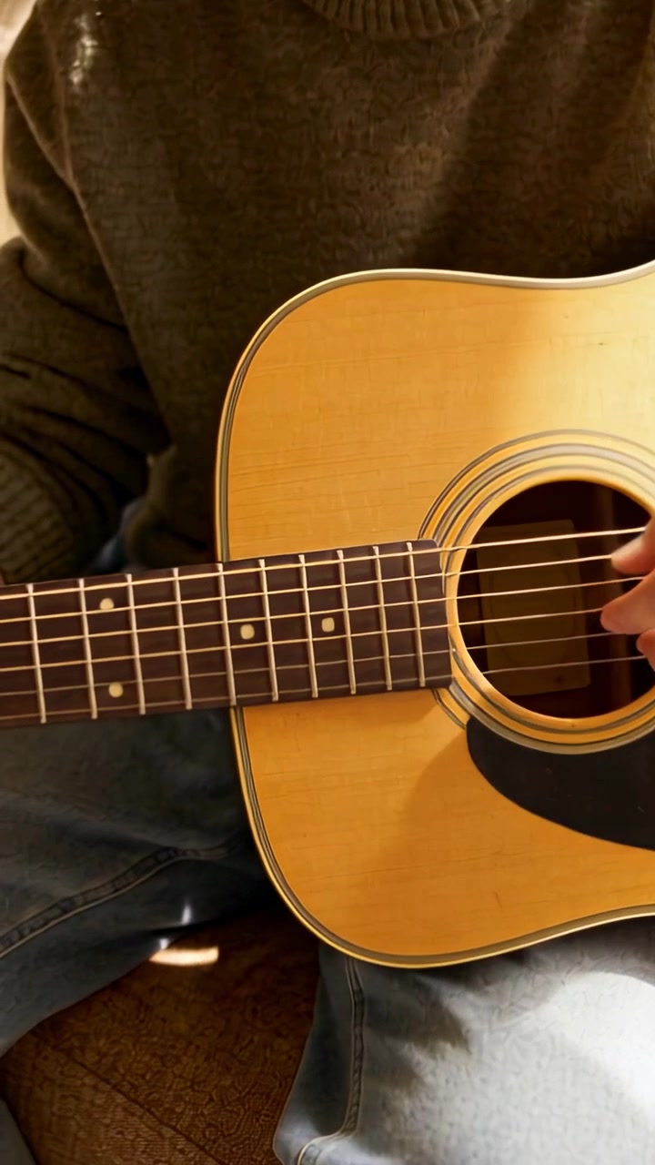Wideo pionowe: Uśmiechnięty mężczyzna w oliwkowym swetrze i dżinsach gra na gitarze akustycznej na sofie, ćwicząc.
