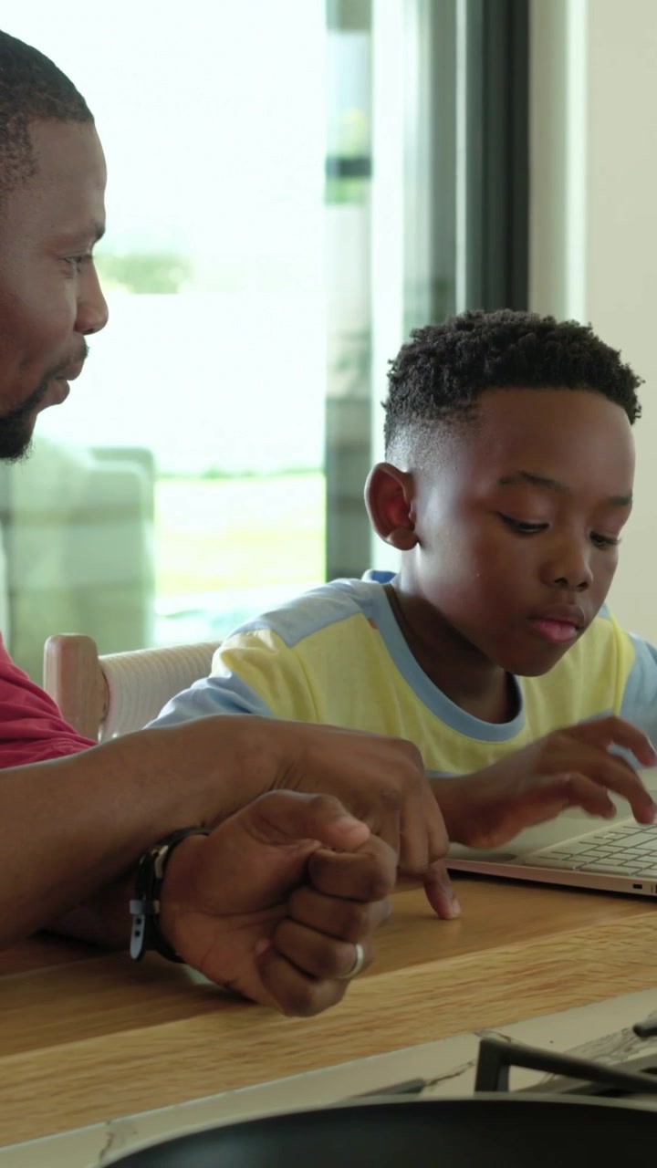 Vertical video: Leaning dad in red guiding son in kitchen pointing as son typing laptop to learn