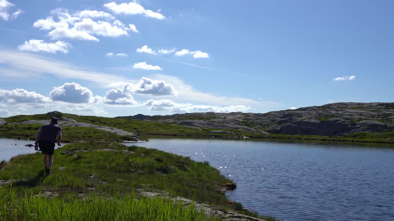 Mann går langs vakker fjellsjø med fiskestang – Sommerferie fjelltur med blå himmel bakgrunn