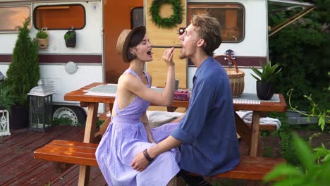 portrait of cute couple man and woman eating raspberries together at wooden table outdoors, feeding each other, kissing. happy vacation together in trailer