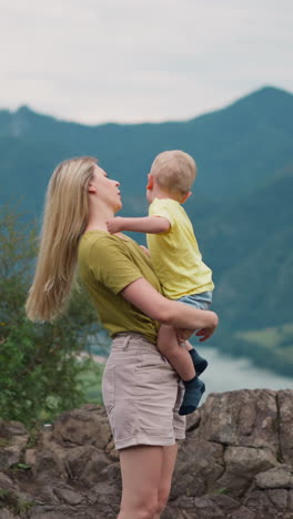 young mother adjusts loose hair holding toddler boy in arms on rocky hill top against mountain silhouettes at highland on gloomy day slow motion