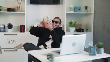 the young serious man in sunglasses is sitting at a table, scattering money, dollar bills around him, rejoicing in the money he has earned