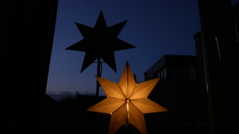 Primer-Plano-De-ángulo-Bajo-De-La-Decoración-De-Estrellas-De-Iluminación-En-La-Ventana-Con-Cielo-Azul-Durante-La-Noche-En-Segundo-Plano.