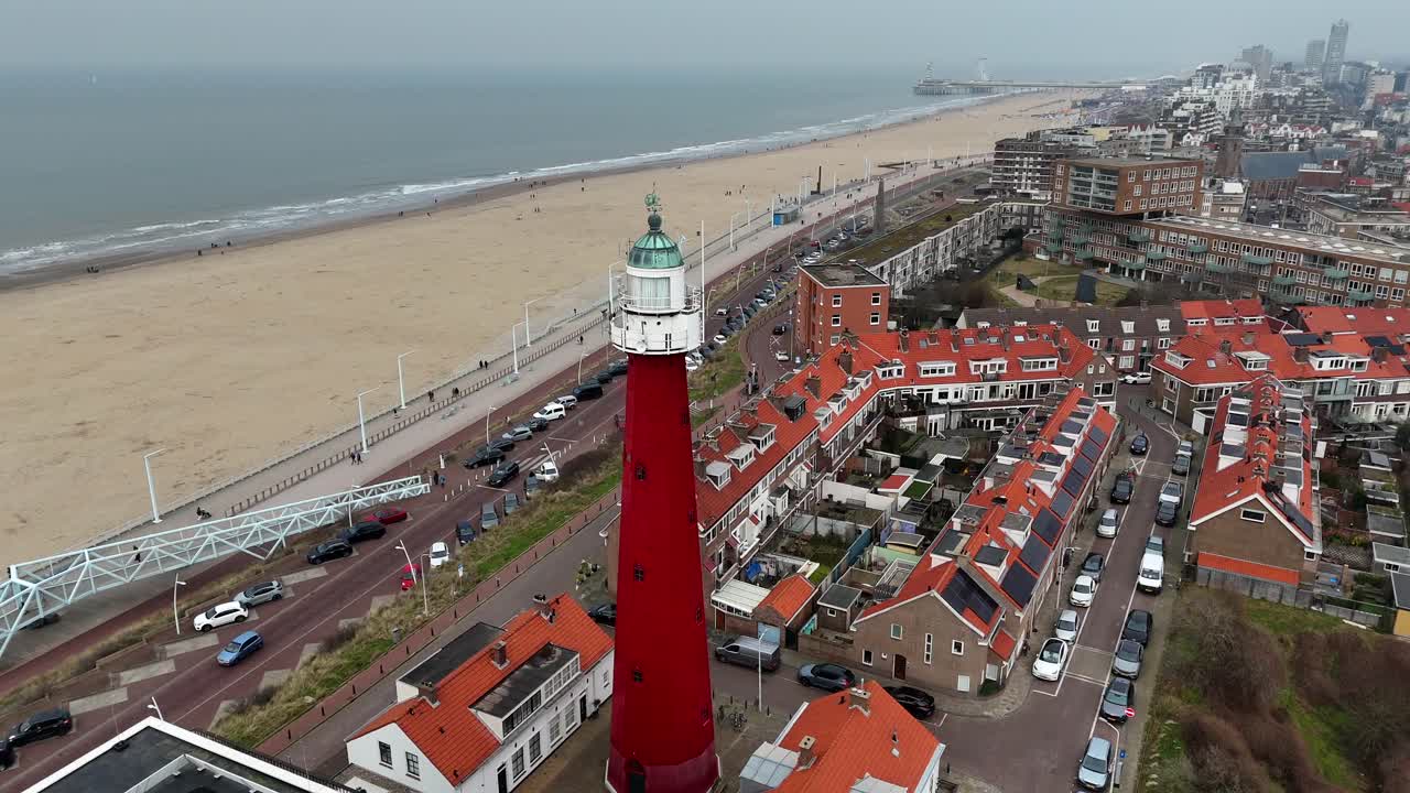 Red lighthouse of Scheveningen Town with sandy beach during cloudy winter day. Aerial orbit shot. Pier and port with housing area in Netherlands near The Hague. Wide shot.