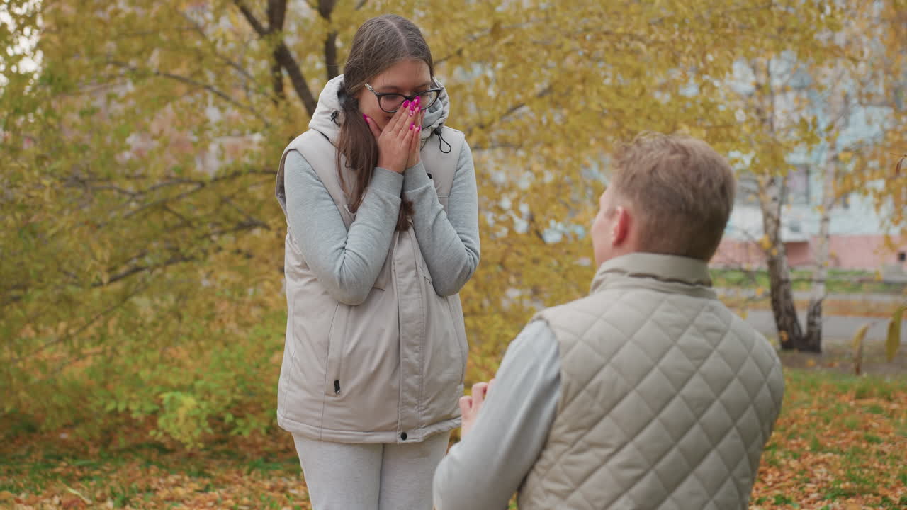 Back view of man kneeling to propose as excited woman with painted nails covers mouth smiling and nodding yes surrounded by autumn leaves with residential building