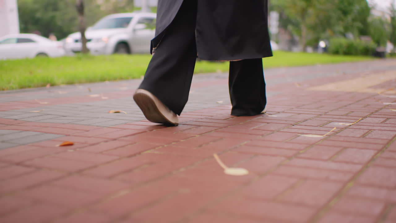 Rear view of woman walking along park pathway in long black coat and white sneakers, focus on steps against brick sidewalk with autumn leaves, conveying movement, solitude, lifestyle, travel atmosphere