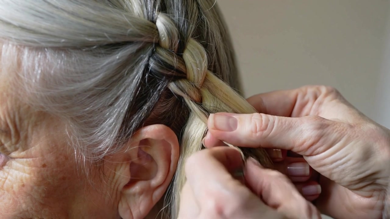 Mani che iniziano a intrecciare una treccia francese a casa, curando il cuoio capelluto di una donna e stringendo i capelli grigi.