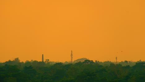 Chimenea-Y-Torre-De-Red-Silueteadas-Contra-El-Vibrante-Horizonte-Naranja-De-La-Puesta-De-Sol-De-Bangladesh-Con-Fuerte-Smog-De-Contaminación