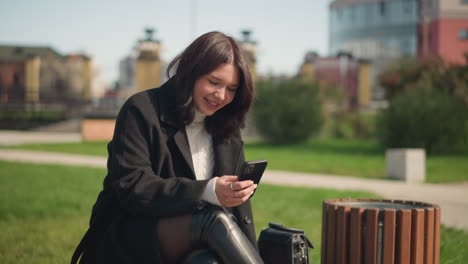 cheerful white lady smiling while using her smartphone in a park, sitting with crossed legs, surrounded by greenery, blurred background featuring a building, enjoying her outdoor moment