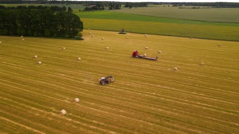 drone-shot-of-a-tractor-working-with-a-cargo-truck-and-hay-rolls-in-a-plain-yellow-field,-tractor-putting-hay-rolls-on-the-cargo-trailer