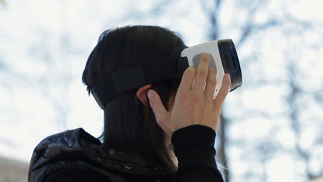 vista lateral de una joven de cabello oscuro con un abrigo negro cálido con gafas de realidad virtual en el parque de principios de otoño