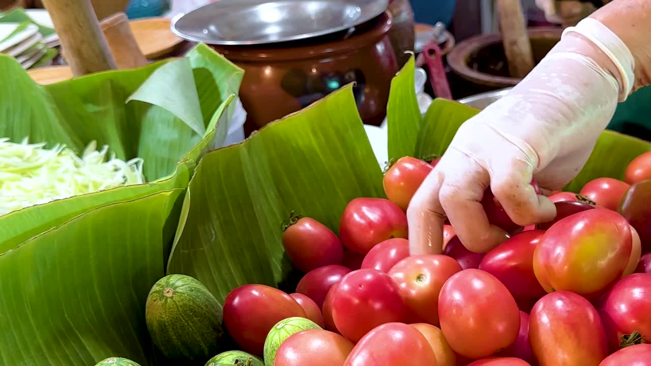 A vendor's gloved hand picks ripe tomatoes from a banana leaf display at a vibrant market stall.