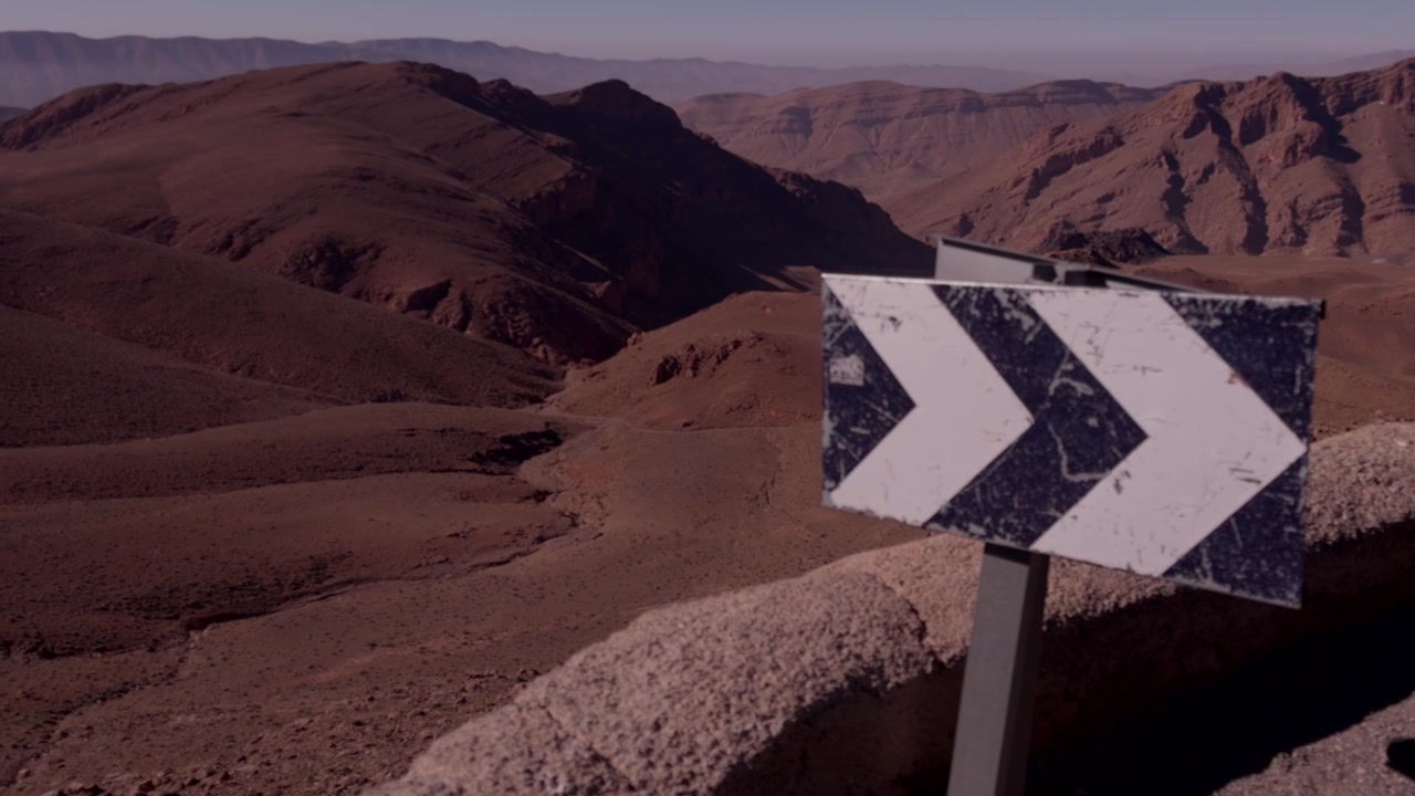 Worn blue and white chevron direction sign on a mountain road edge above a vast arid desert valley