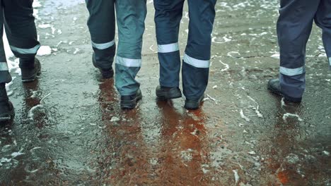four workers in a gray uniform with a horizontal white stripe on their pants are walking along the asphalt with wet snow in black boots. close-up of legs.