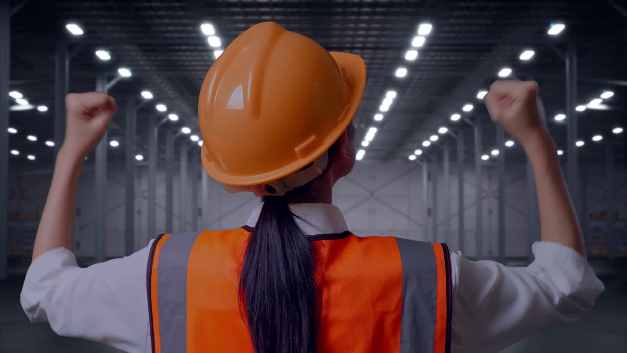 Vista ravvicinata da dietro di un'ingegnera con casco di sicurezza che alza le mani in segno di celebrazione mentre lavora in un magazzino moderno e vuoto.