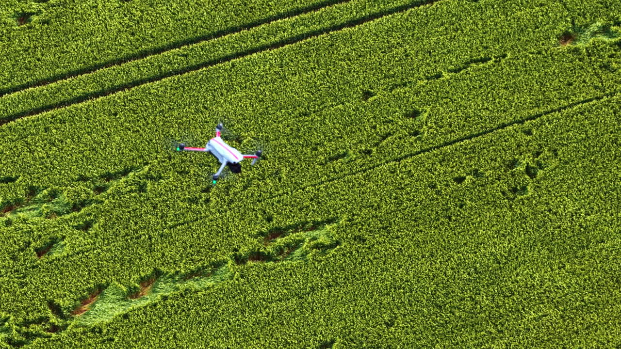 A top-down aerial tracking shot follows a quadcopter drone as it flies over a vast green agricultural field, used for modern precision farming and surveillance