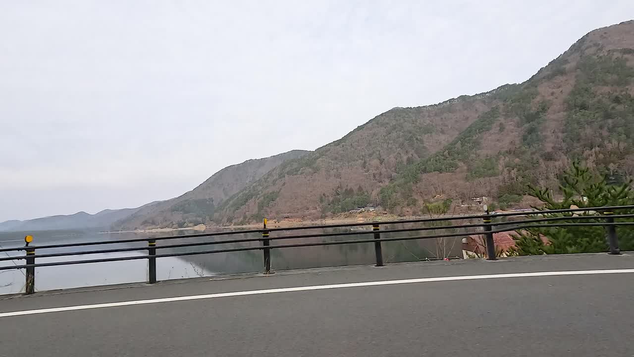 A serene drive along Lake Kawaguchi with mountain views, captured in soft daylight, showcasing the tranquil beauty of Fujikawaguchiko