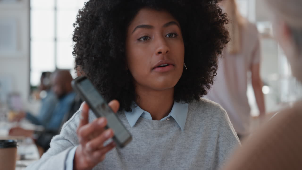 young mixed race business woman manager using smartphone training intern pointing at screen sharing feedback discussing ideas with colleague in office