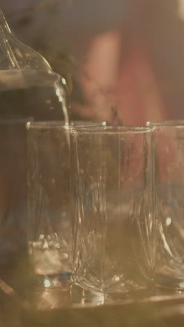primer plano de alguien vertiendo agua infundida de una jarra en tres vasos bajo la cálida luz del sol, rebanadas de naranja y hojas de menta visibles en el agua, con fondo borroso