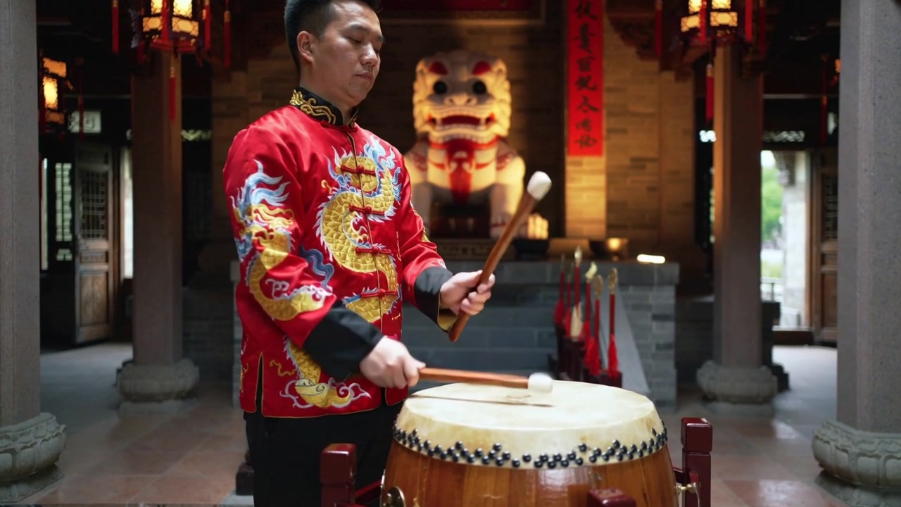 Músico com jaqueta vermelha erguendo baquetas, tocando tambor de barril em templo, ao lado de leão guardião, para ritual.