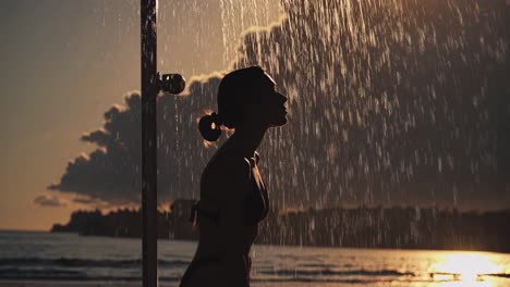 Woman enjoys a refreshing beach shower as the sun sets in the background, creating a stunning silhouette. The scene captures tranquility and the beauty of nature at dusk