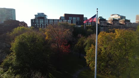 Vista-Aérea-Levantando-La-Bandera-Nacional-Estadounidense-Ondeando-En-El-Jardín-Público-De-Boston-En-La-Mañana-Ventosa