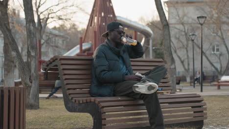 side view of dark skinned man wearing cap and sunglasses sitting on park bench holding sandwich and bottle of juice during outdoor break in urban park surrounded by bare trees on calm afternoon