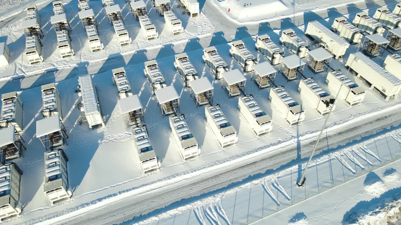 Luftfoto, der bevæger sig forbi rækker af store batterilagringsenheder i netværksstørrelse, der opererer under klare vinterforhold og understøtter en pålidelig elforsyning. Massiv BESS-installation, batteripark-layout.