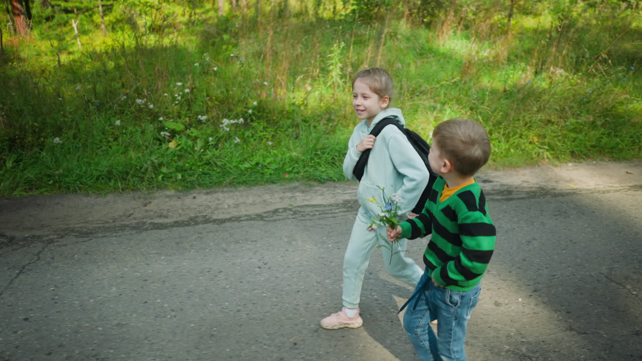 Young girl adjusts bag strap while walking and laughing with younger brother holding wildflowers on paved forest path surrounded by tall green grass and trees under daylight atmosphere