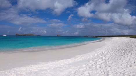 a white sand beach with turquoise and clear water of a paradisiac island of the galapagos islands. there are cruises and some islands on the clear and big ocean.