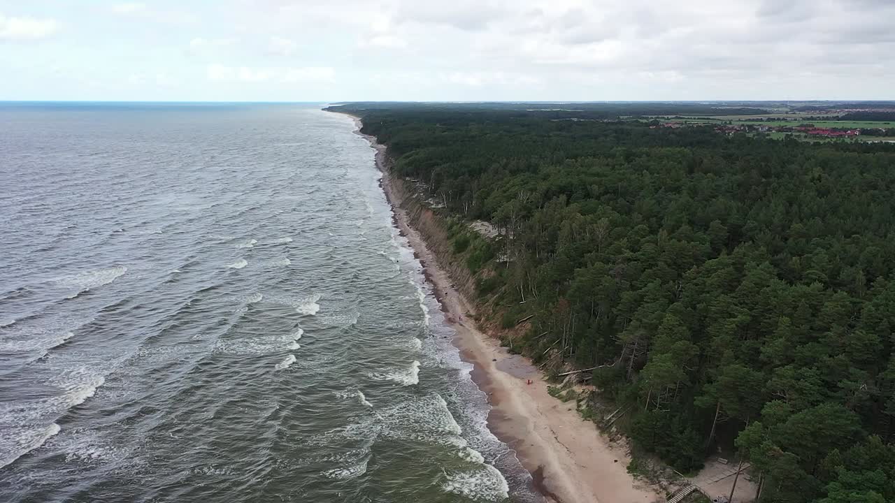 Revealing a majestic cliff on the Baltic Sea coastline in Lithuania through drone footage, the scene unfolds with tumultuous waves and the vivid green tones of the water under moody skies