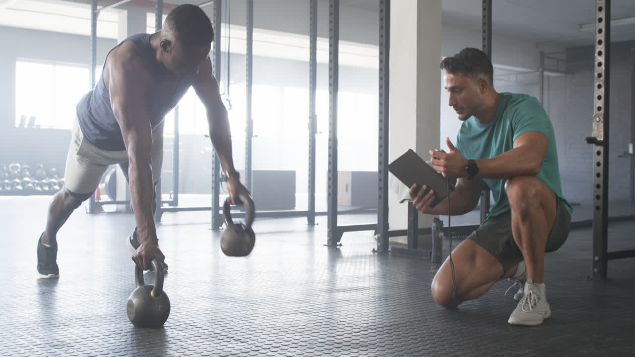 Strong, fit biracial man trains with kettlebells while young trainer holds a tablet