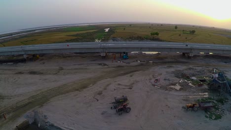 Aerial-view-of-Under-constructed-bridge-over-the-river-with-heavy-duty-trucks-and-machinery,-Farms-in-the-river