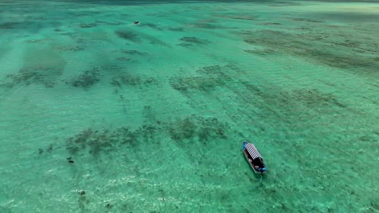 Zanzibar'da kristal berraklığında turkuaz sularda yüzen bir teknenin huzurlu sinematik görüntüsü. Sakin okyanus, canlı mavi-yeşil tonlar ve dingin ada atmosferiyle çarpıcı tropikal manzara.