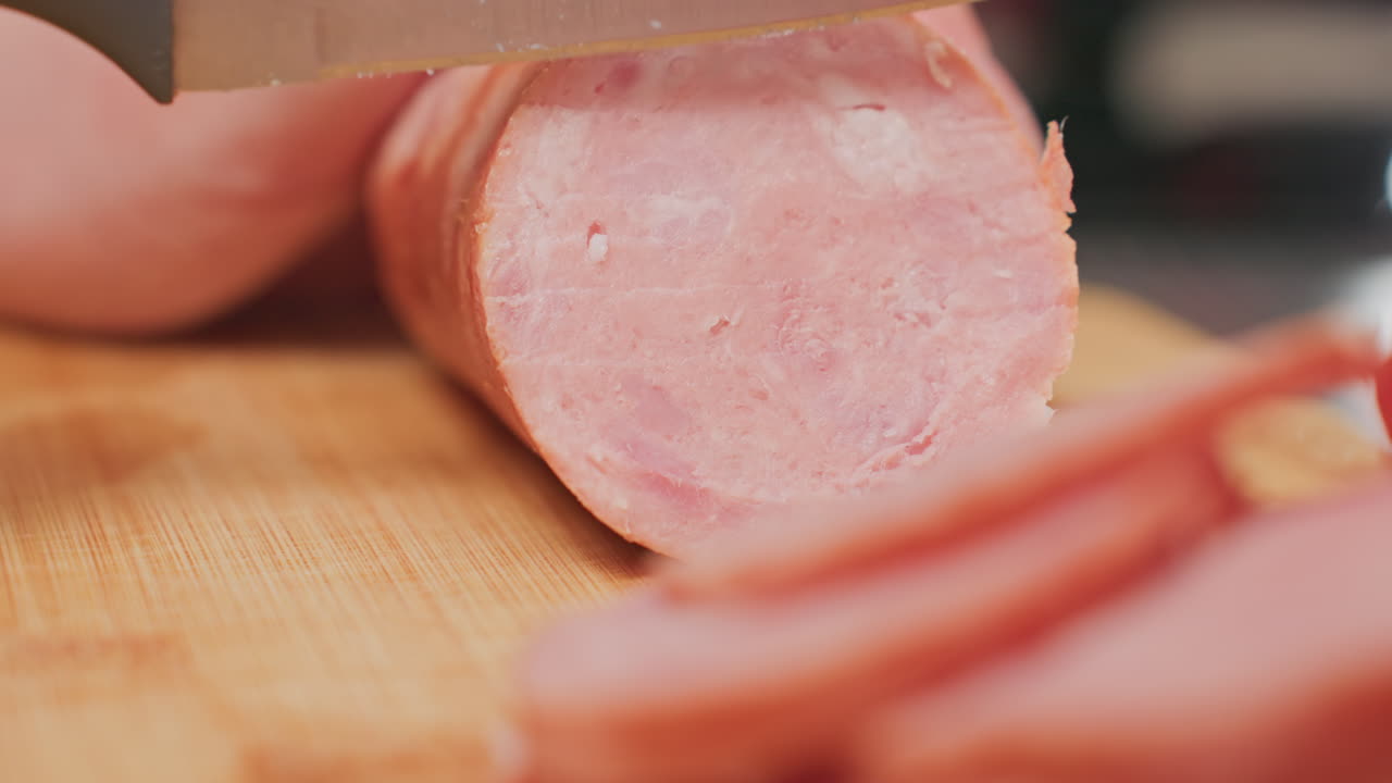 close up person slicing round pink meat sausage with sharp kitchen knife on wooden cutting board showing smooth clean cuts, juicy texture, and preparation process