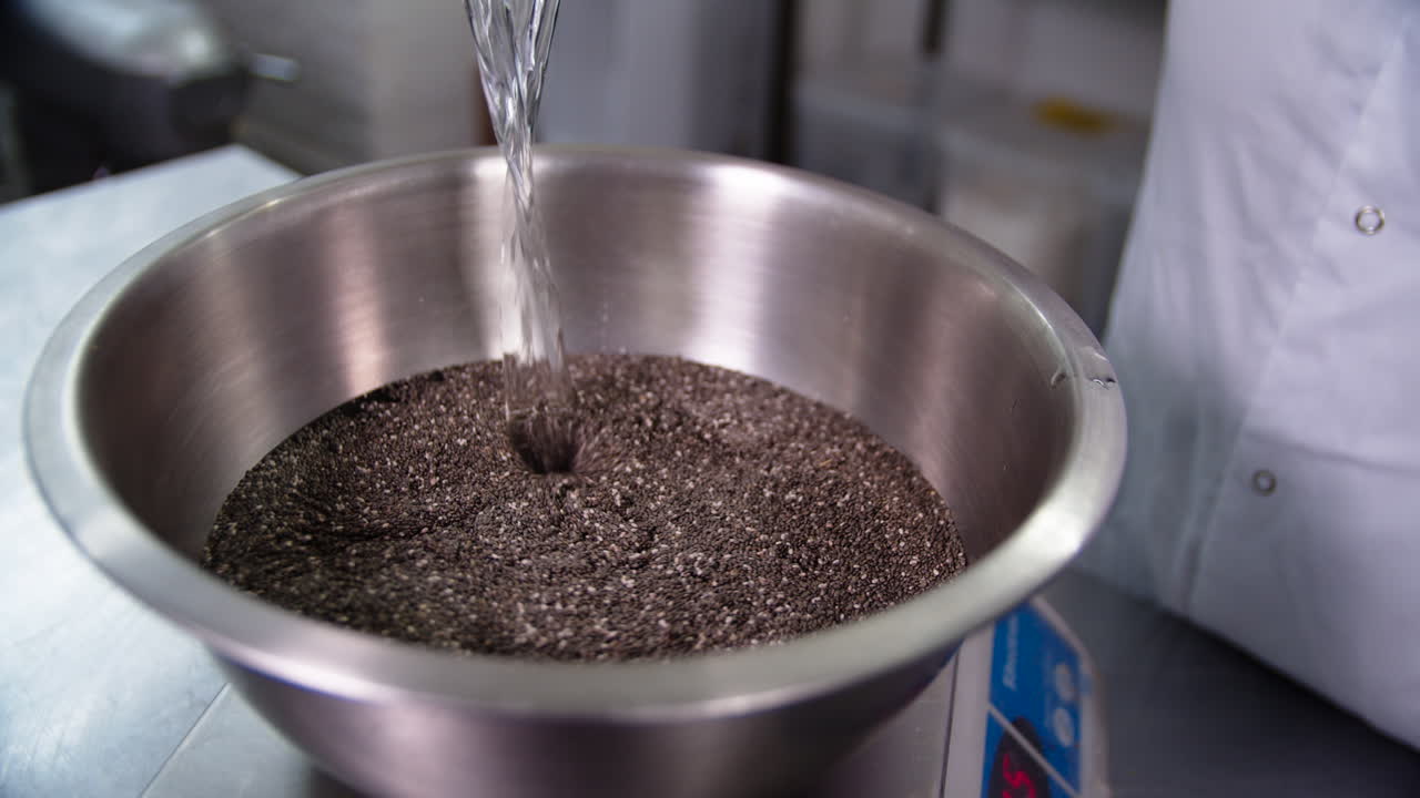 Una grande ciotola metallica piena di semi di chia, su cui una chef in divisa bianca versa dell'acqua