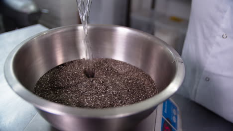 a large metallic bowl full of chia seeds has water poured on it by a female chef in kitchen whites