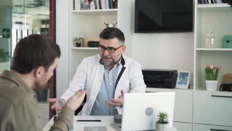 the doctor sits at the desk in a bright modern office and talks to his patient. the patient is saddened by the conversation with the doctor.