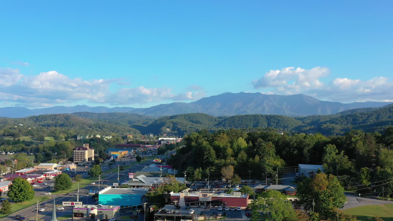 Breathtaking aerial panorama of a Tennessee town framed by the rugged Great Smoky Mountains under a brilliant blue sky with fluffy clouds. Captures the essence of mountain living and bustling streets