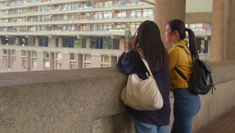 vista trasera de dos jóvenes amigas que visitan el centro barbican en la ciudad de londres tomando fotos en el teléfono móvil 2