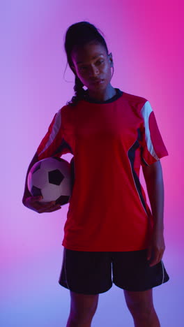 Vertical-Video-Studio-Portrait-Of-Female-Football-Or-Soccer-Player-Wearing-Team-Shirt-Holding-Ball-Under-Her-Arm-Shot-Low-Key-Against-Colourful-Mixed-Lighting