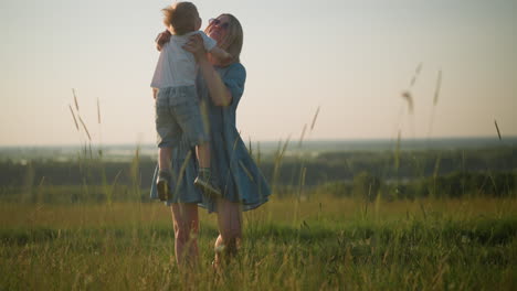 a woman in a blue dress stands in a grassy field, warmly embracing a little boy in white as he jumps into her arms. the scene captures a tender moment of love and connection between mother and child