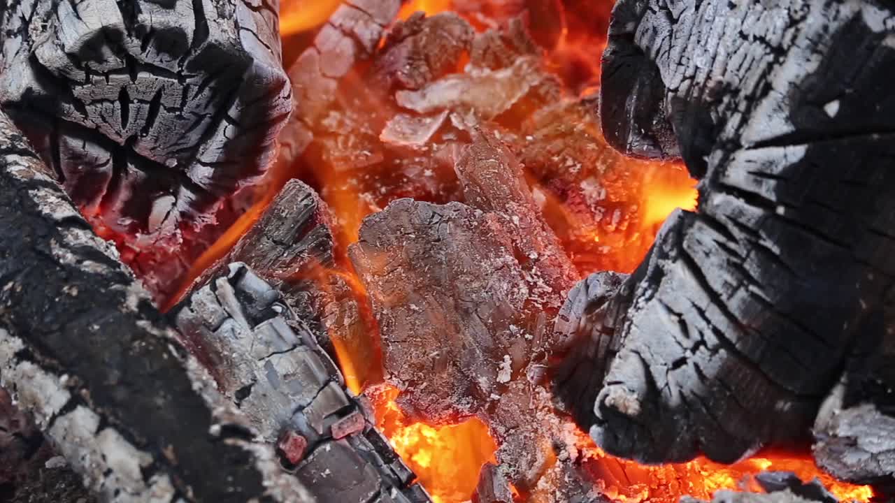 Closeup shot of glowing campfire embers with burning logs showing bright orange flames, charred wood texture, and detailed ash, capturing warmth and rustic outdoor fire atmosphere