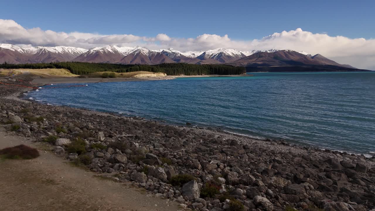 4K view of the rocky shoreline of Lake Pukaki. Blue water contrasts with the rugged snow-capped Southern Alps in the background. A stunning landscape in New Zealand's South Island