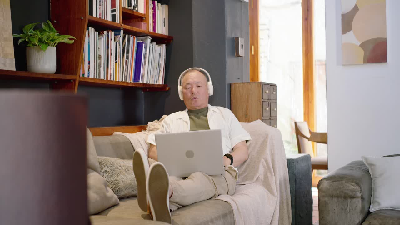 Camera shifting from pipe, Senior Asian man reclining on sofa, working on laptop wearing headphones